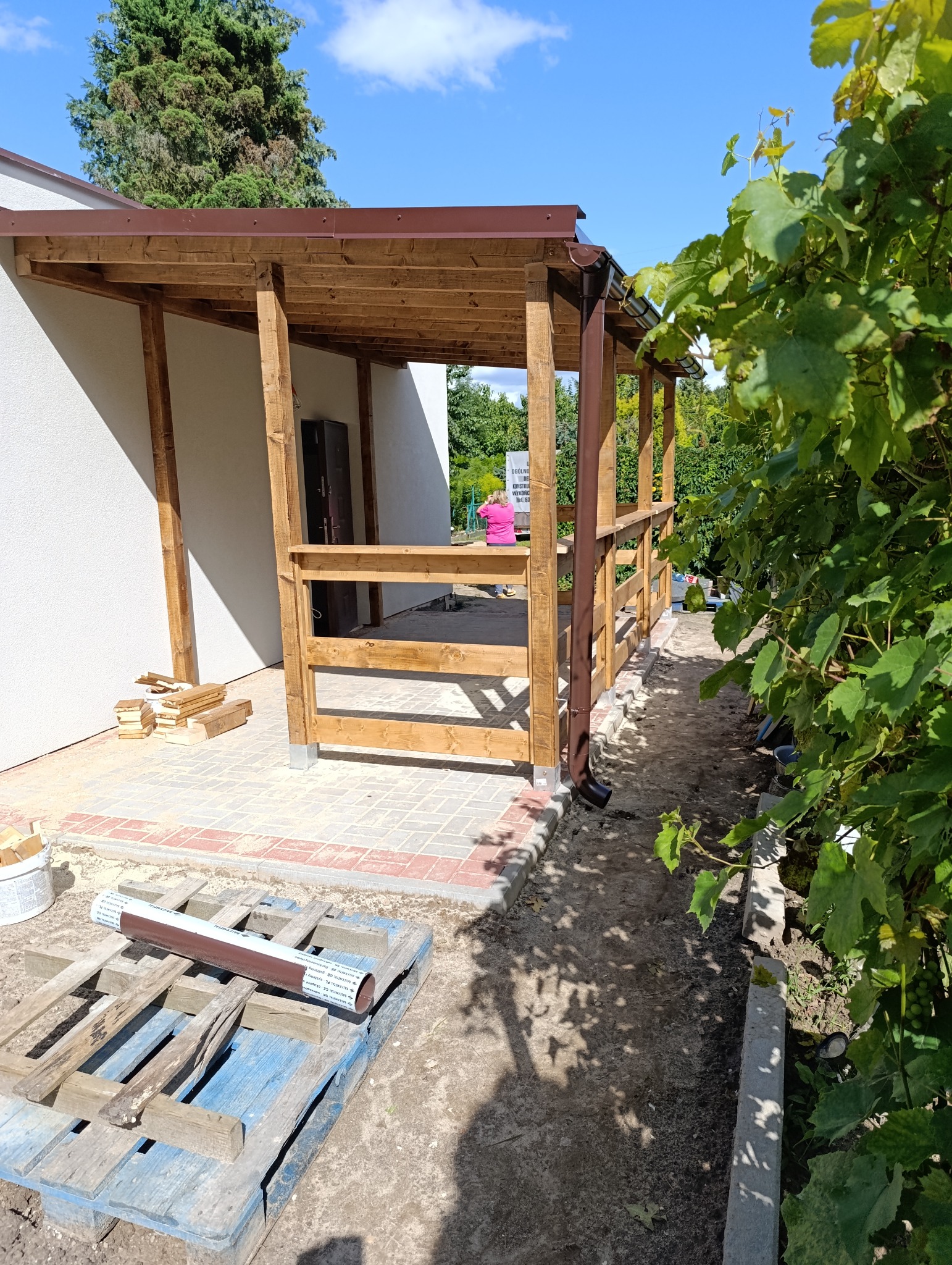 Drewniana, zadaszona weranda w trakcie budowy. Widoczne elementy konstrukcyjne, brązowe rynny, paleta z materiałami i fragment ogrodu z winoroślą.