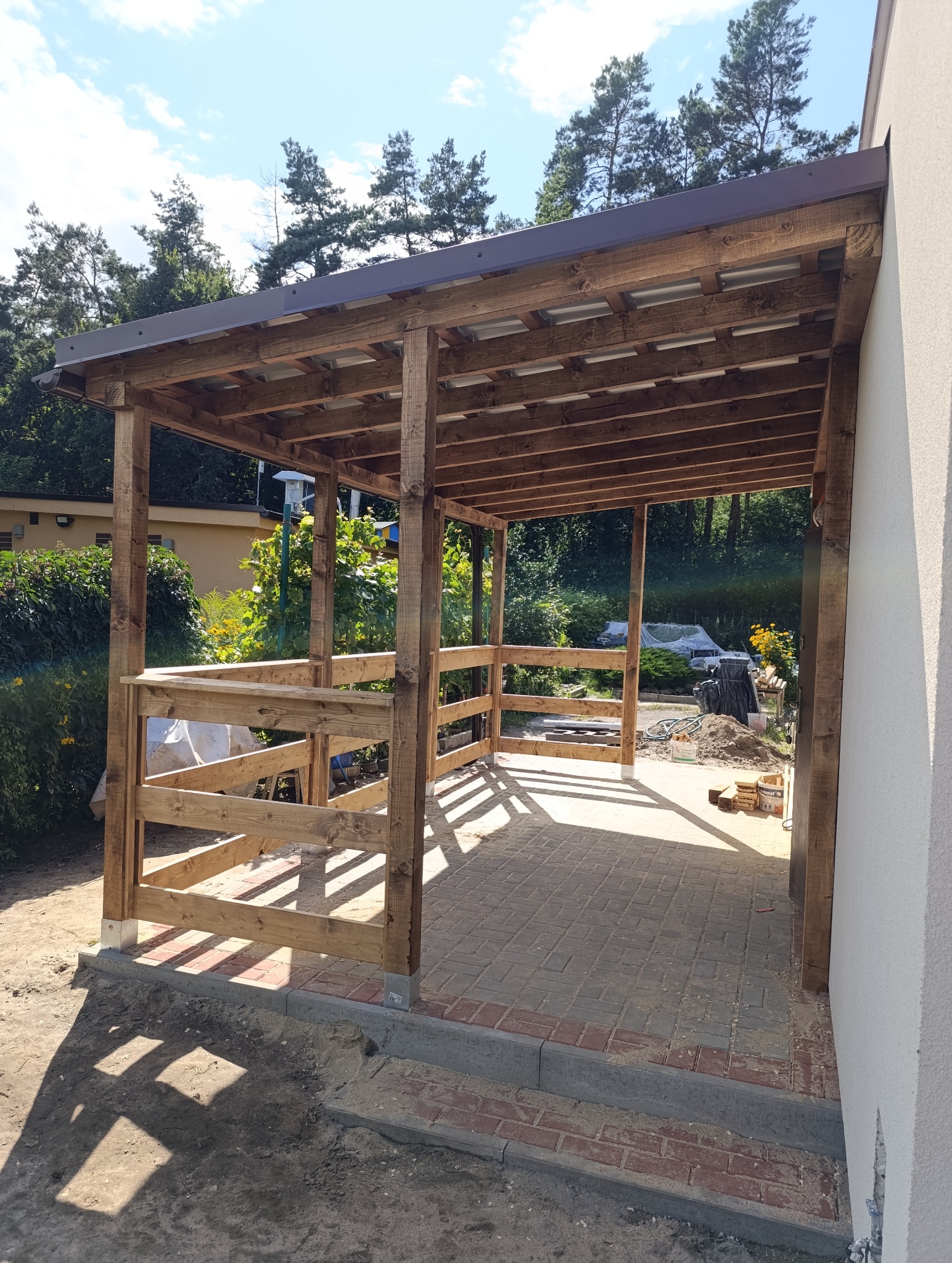 Drewniana konstrukcja z zadaszeniem, prawdopodobnie altana lub wiata, w trakcie budowy. Widoczne elementy konstrukcyjne, drewniane słupy i zadaszenie z blachy.