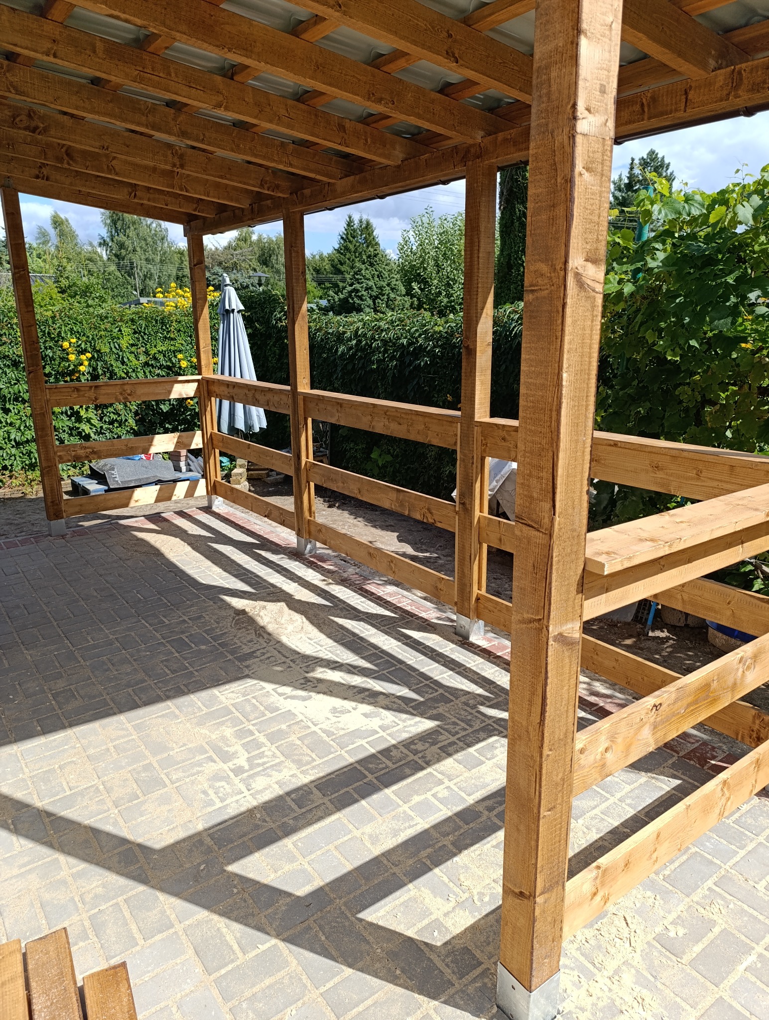 Drewniana konstrukcja zadaszenia tarasu w trakcie budowy, widoczne słupy, balustrady i zadaszenie. W tle zieleń ogrodu i parasol. Cień rzucany przez konstrukcję na kostkę brukową.