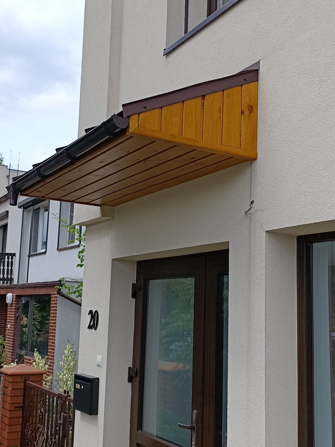Drewniana, zadaszona weranda nad brązowymi drzwiami wejściowymi do domu z numerem 20. Widoczna czarna skrzynka na listy i fragment sąsiedniego budynku.