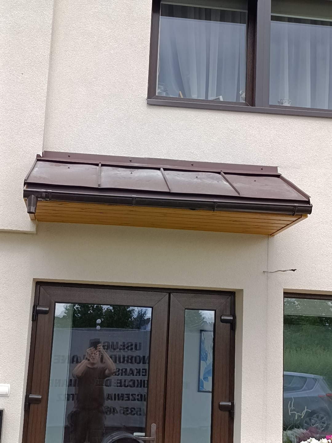 Brązowy daszek nad wejściem do budynku z brązowymi oknami. Widoczne odbicie osoby robiącej zdjęcie w szybie drzwi.
