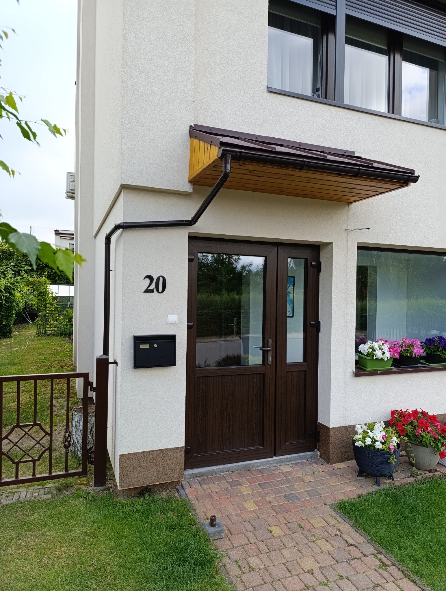 Elewacja domu z numerem 20, brązowe drzwi wejściowe pod zadaszeniem, skrzynka pocztowa, kwiaty w donicach przy oknie, kostka brukowa i trawnik.