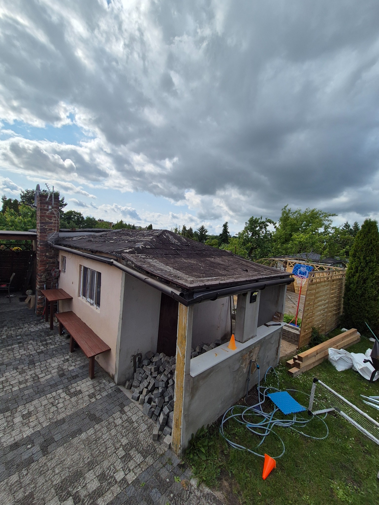 Przygotowania do wymiany dachu na budynku gospodarczym z kominem. Stare pokrycie dachowe, obok kostka brukowa i materiały budowlane.