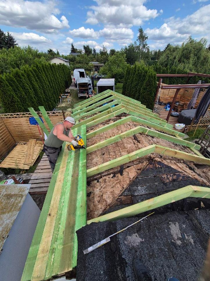 Prace dekarskie: mężczyzna w trakcie montażu drewnianej konstrukcji dachu, widoczne ocieplenie i narzędzia. W tle zieleń drzew i zabudowania.