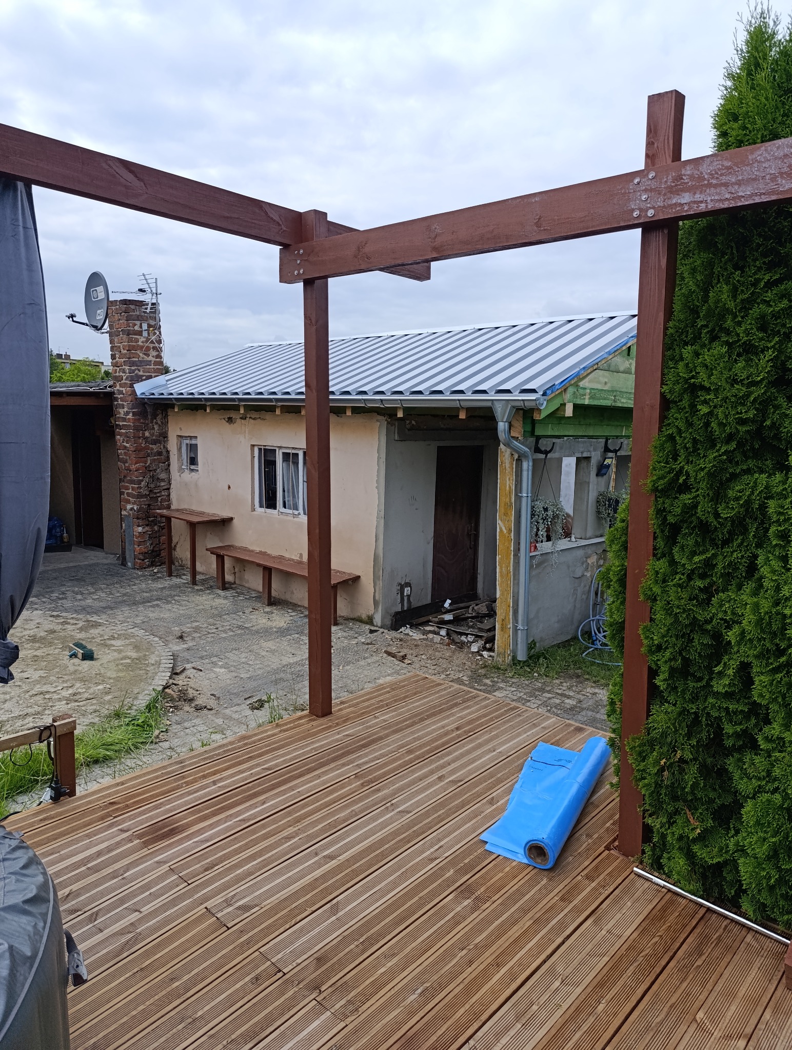 Nowy, blaszany dach z orynnowaniem na budynku gospodarczym, widok z drewnianego tarasu z pergolą. Częściowo zdemontowane elementy konstrukcji.