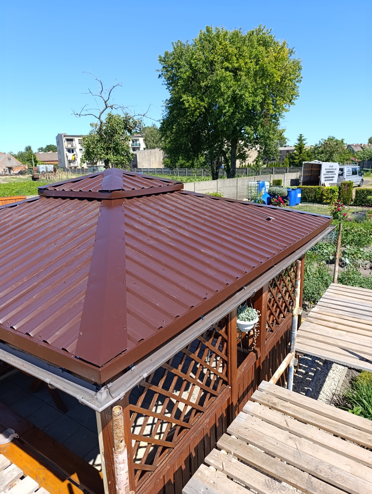 Nowy, brązowy dach z blachy trapezowej na altanie ogrodowej z drewnianą konstrukcją kratową. Widoczne rynny i fragment ogrodu w tle.