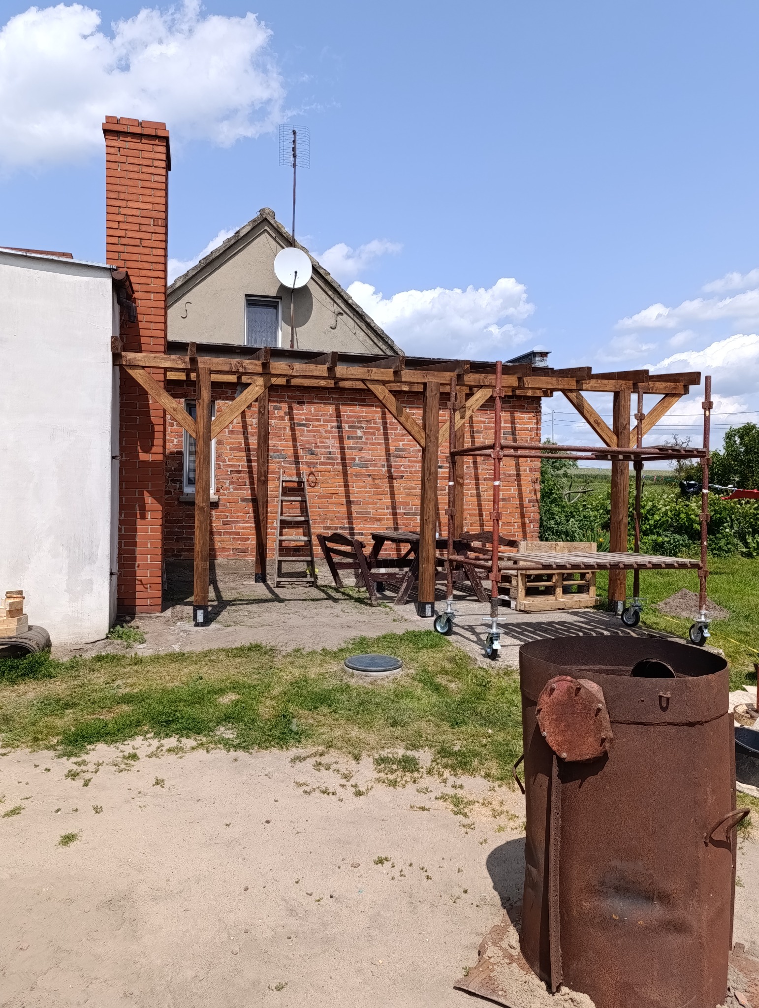 Drewniana konstrukcja zadaszenia tarasu przy ceglanym domu, widoczne rusztowanie na kółkach i metalowy piec. Słoneczny dzień, błękitne niebo z chmurami.