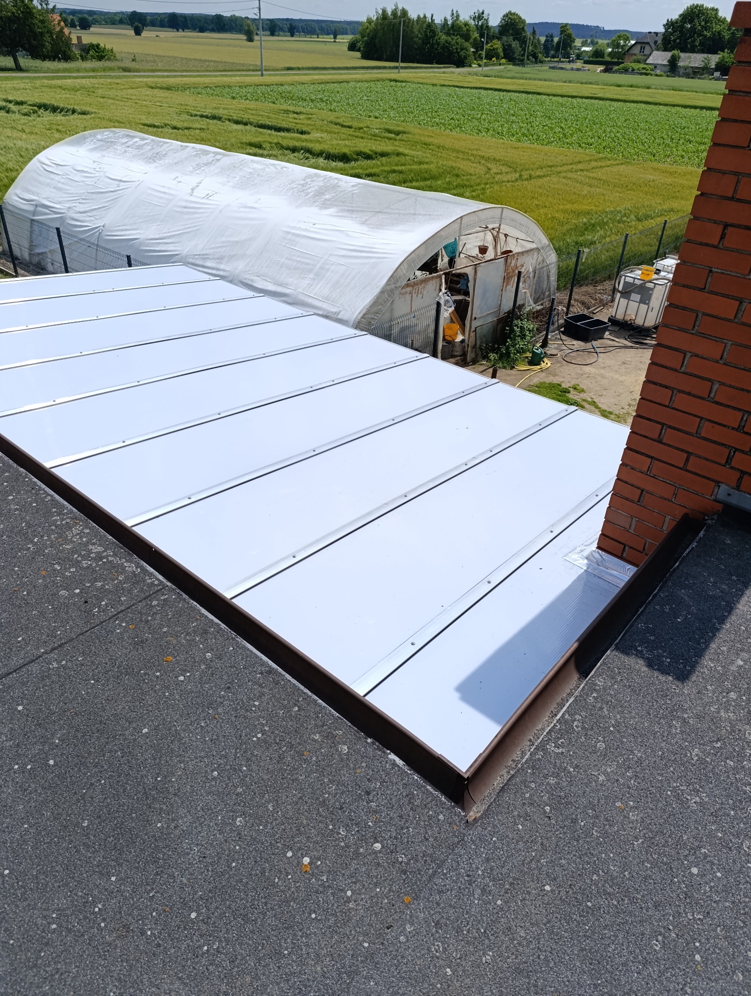 Nowe, białe panele dachowe z metalowymi listwami na tle zielonego pola i szklarni. Widoczny fragment komina z cegły i pokrycie dachu z papy.