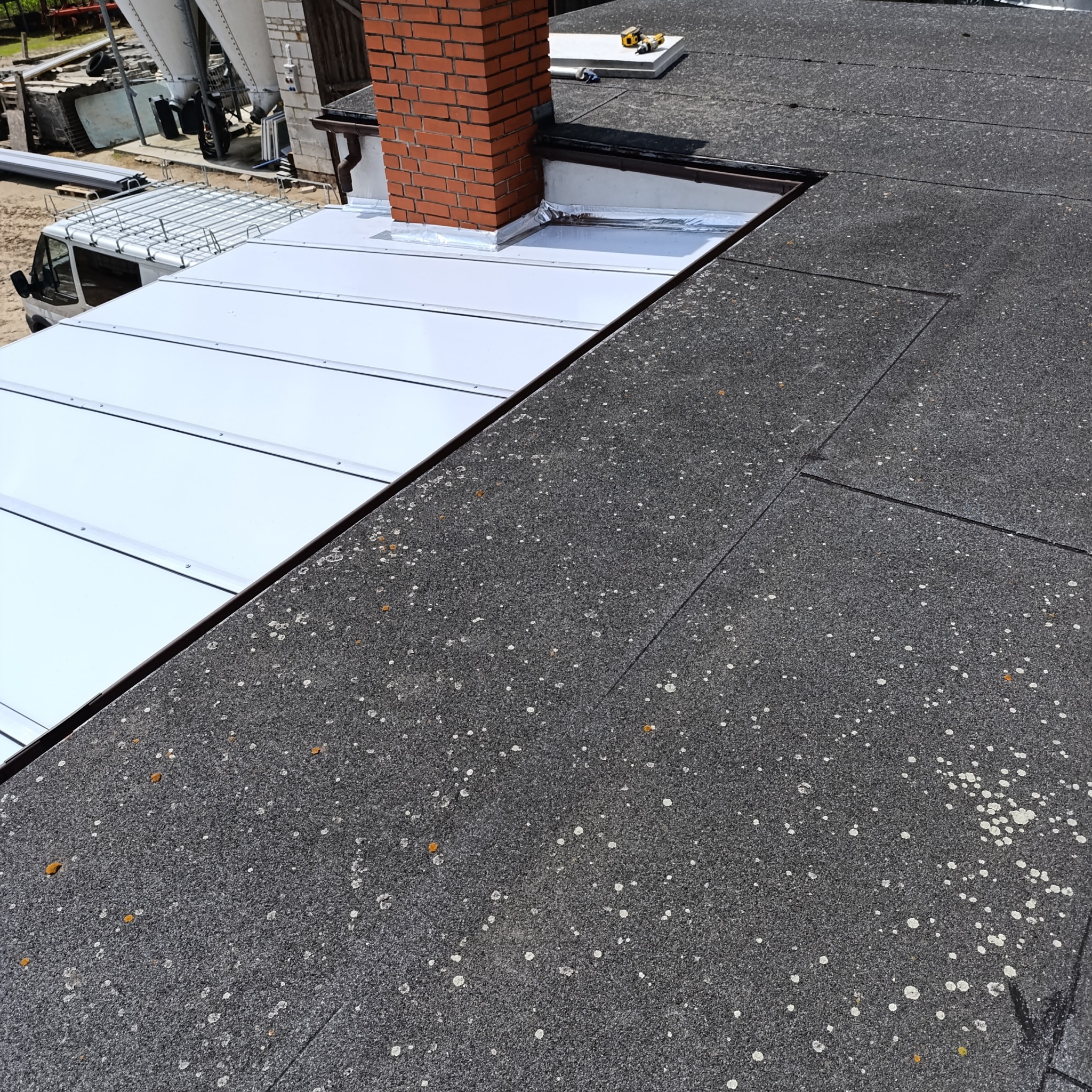 Fragment dachu: stara, bitumiczna papa z plamami i nowa, metalowa część obok komina z cegły. Widoczny samochód dostawczy z elementami dachu.