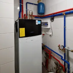 Nowoczesna instalacja pompy ciepła Daikin w kotłowni z białymi płytkami, widoczne rury w izolacji czerwonej i niebieskiej, manometry, zbiornik wyrównawczy i skrzynka elektryczna.