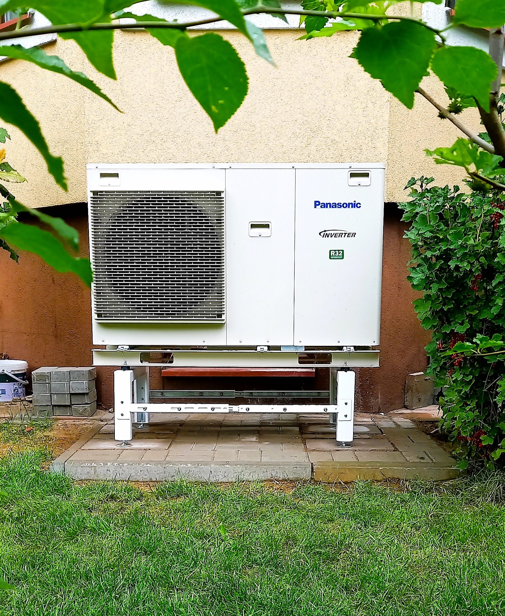 Zewnętrzna jednostka pompy ciepła Panasonic Inverter R32 na metalowej konstrukcji, ustawiona na kostce brukowej, z zielonym krzewem po prawej stronie i fragmentem drzewa w górnej części kadru.