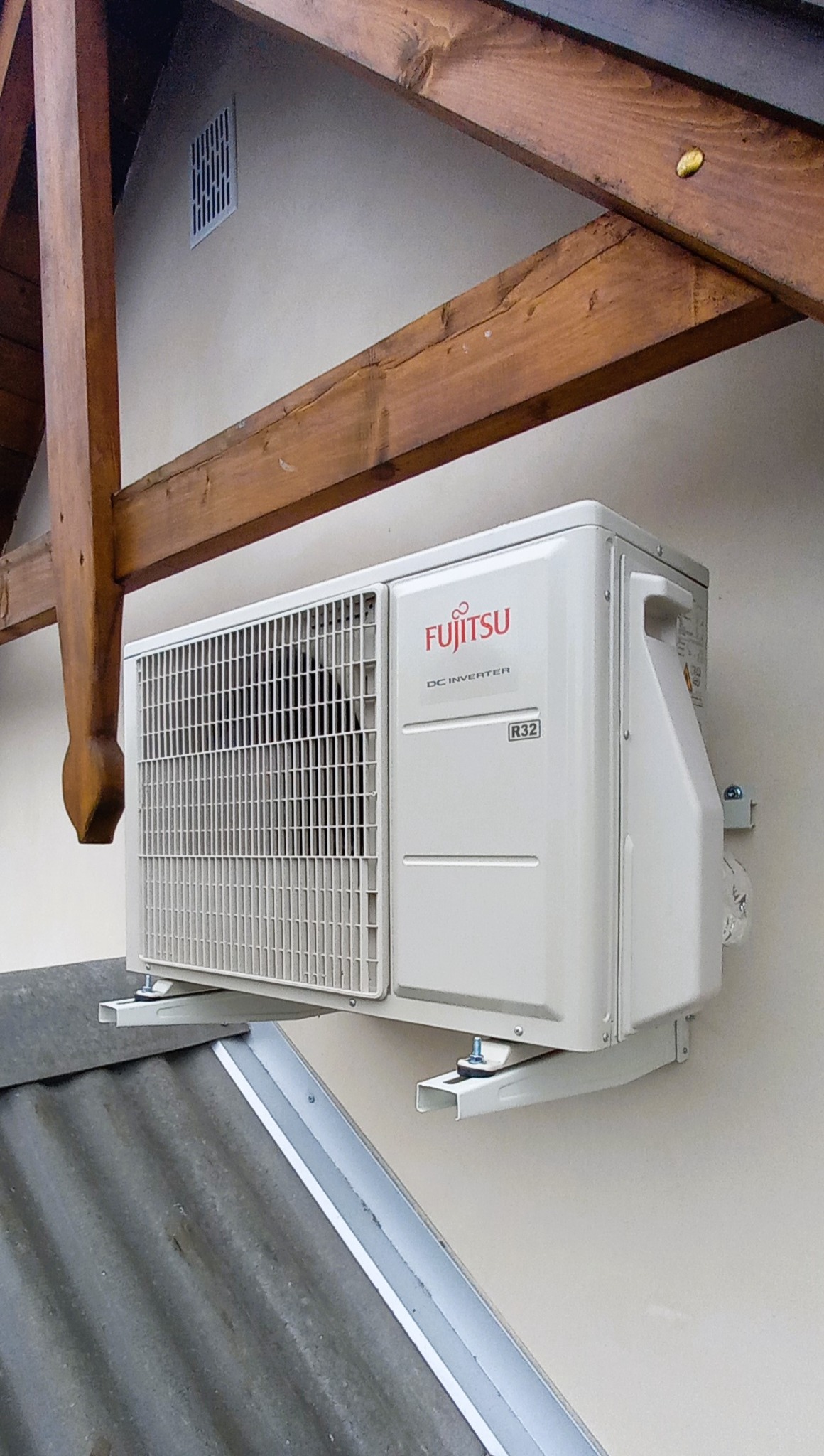 Zewnętrzna jednostka klimatyzacji Fujitsu DC Inverter R32 zamontowana na ścianie budynku, widoczna z boku i od dołu, z fragmentem dachu i drewnianą konstrukcją dachu w tle.