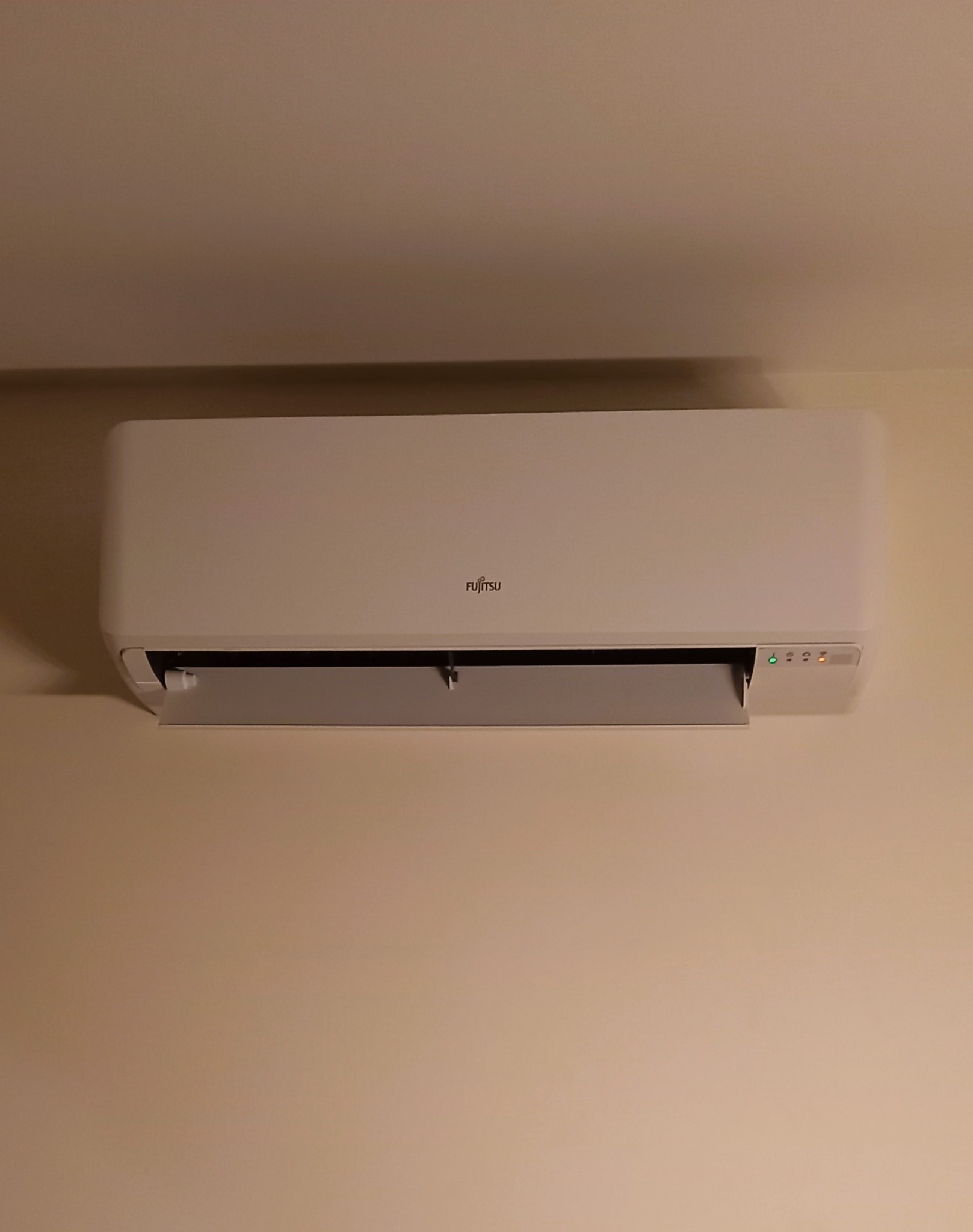 Klimatyzator Fujitsu zamontowany na beżowej ścianie, widoczne diody sygnalizacyjne i otwarty wylot powietrza.