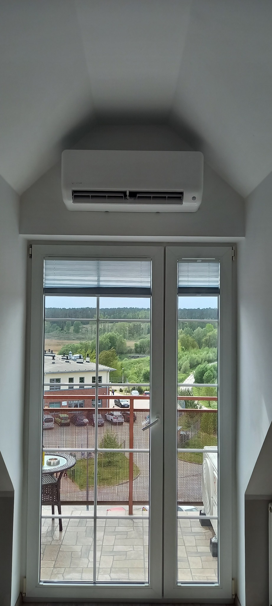 Klimatyzator zamontowany nad dwuskrzydłowymi drzwiami balkonowymi z widokiem na zewnątrz, na balkonie stolik i krzesła.
