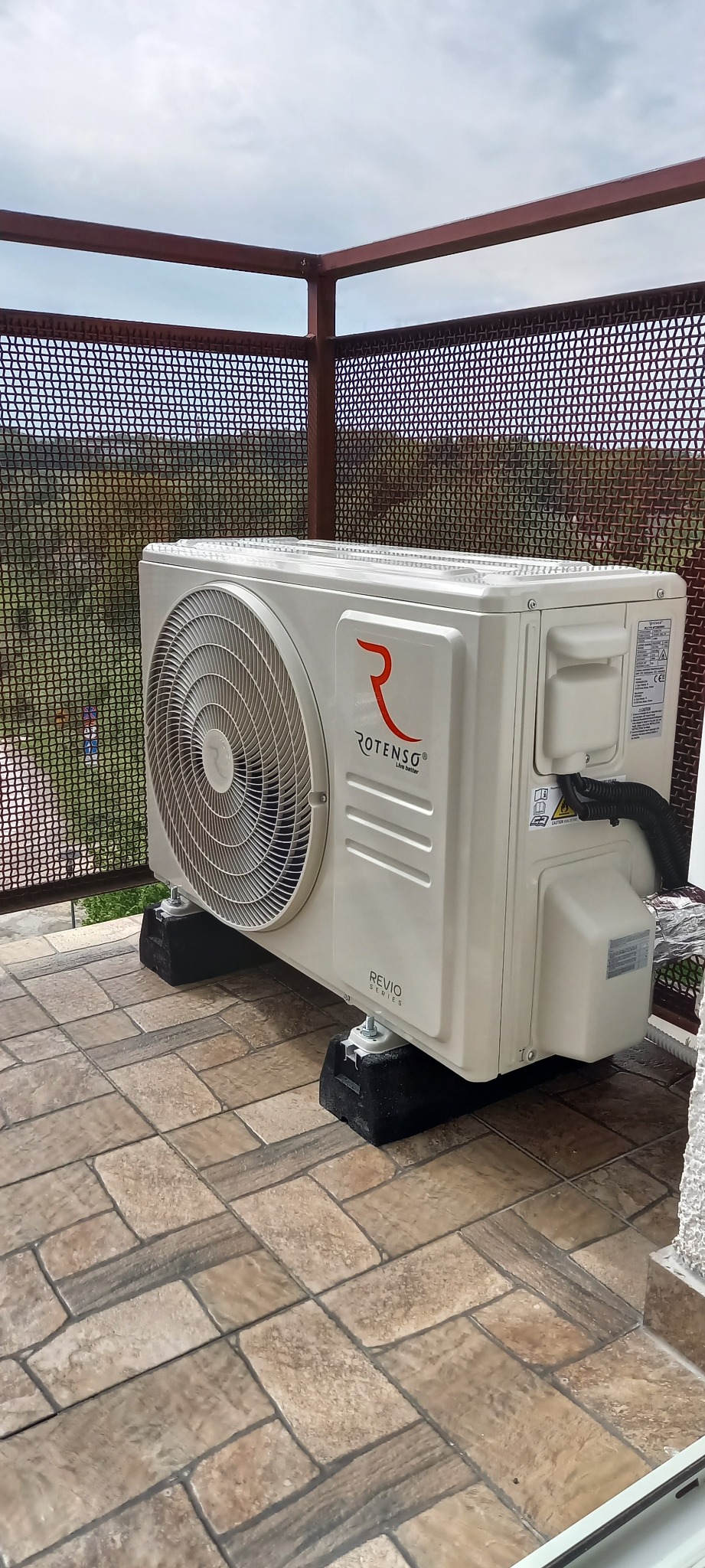 Zewnętrzna jednostka klimatyzacji Rotenso Revio Series na tarasie z widokiem na zalesione wzgórza, zamontowana na gumowych podkładkach antywibracyjnych.