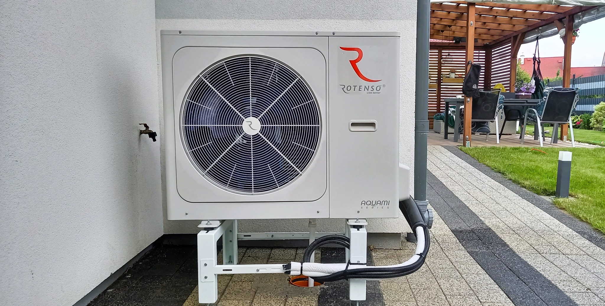 Zewnętrzna jednostka pompy ciepła Rotenso Aquami Series zamontowana na białym wsporniku przy ścianie budynku, z widocznym fragmentem ogrodu i altaną w tle.