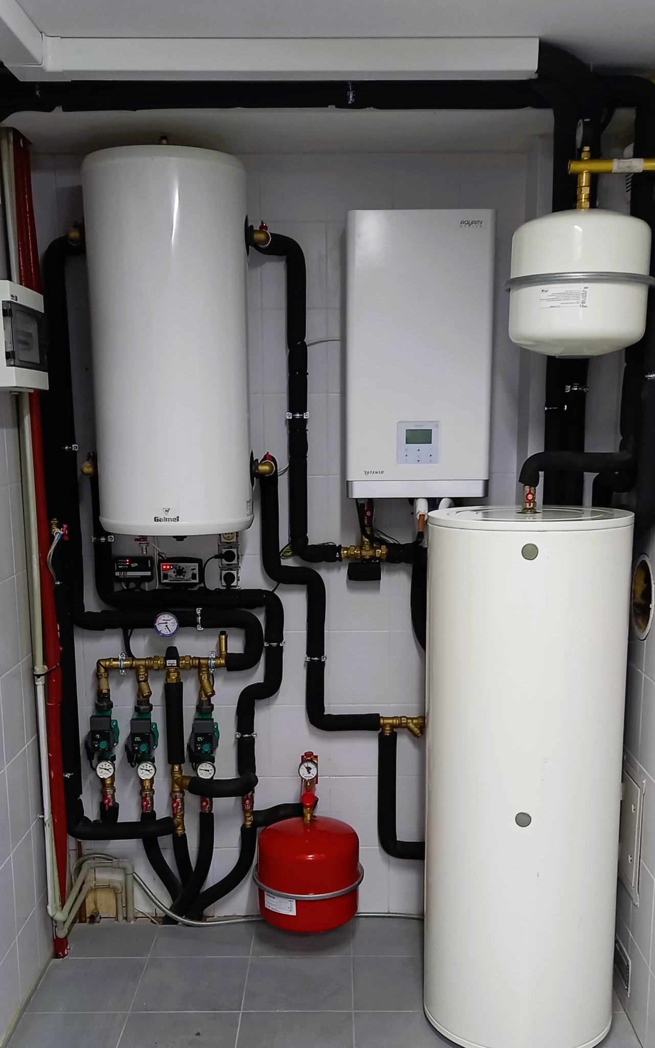 Kompletna instalacja centralnego ogrzewania w kotłowni, widoczne dwa białe zbiorniki akumulacyjne Galmet, biały piec Rotenso, czerwony zbiornik przeponowy, czarne rury izolowane, manometry i zawory