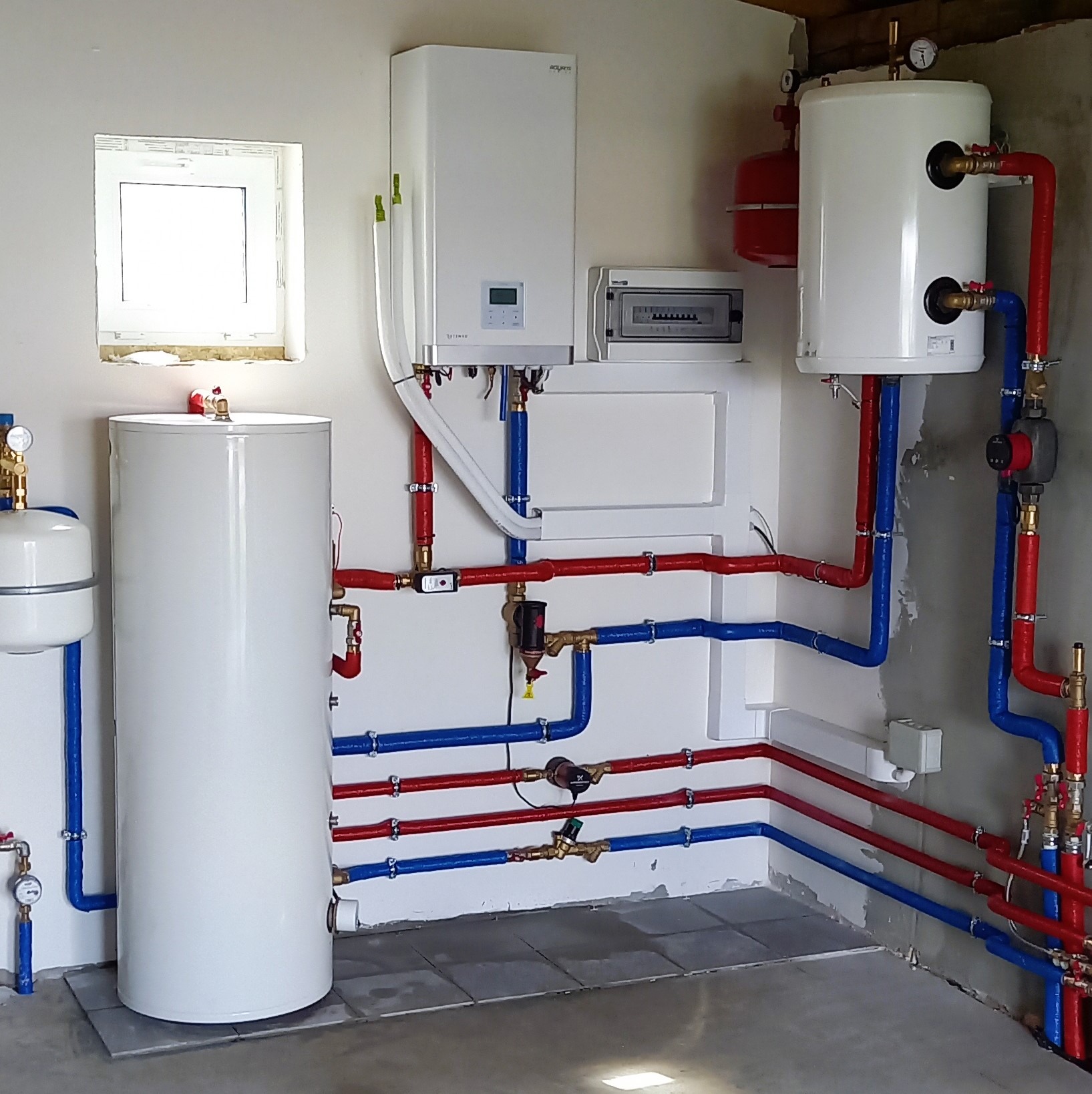 Kotłownia z białym piecem, dwoma zbiornikami, rozdzielnicą elektryczną i instalacją z rur w izolacji czerwonej i niebieskiej, widok na całą instalację z niskiego kąta.