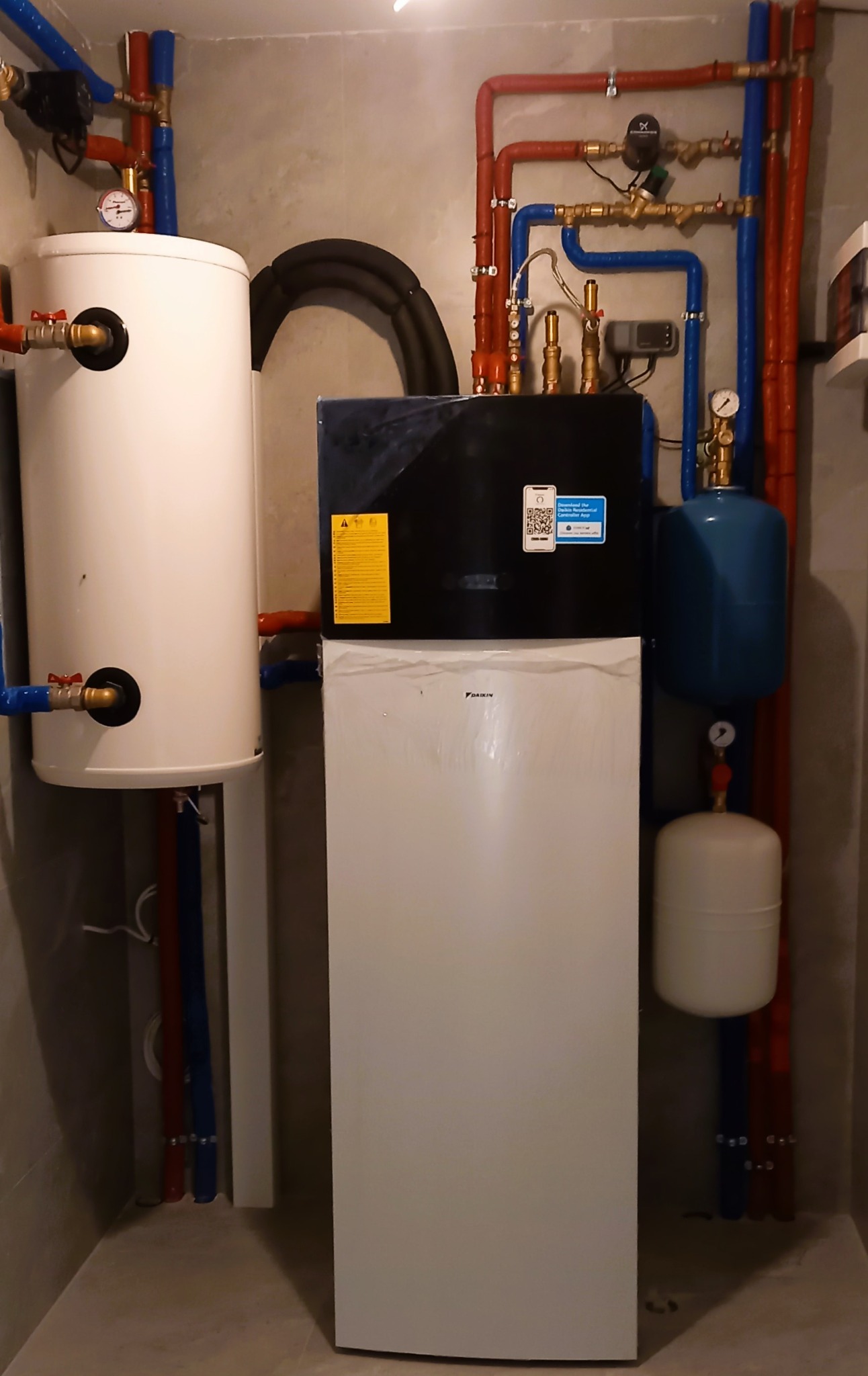 Zestaw instalacji grzewczej z białym zasobnikiem ciepłej wody, niebieskim zbiornikiem wyrównawczym i czarno-białą pompą ciepła Daikin, z widocznymi rurami w kolorze czerwonym i niebieskim...