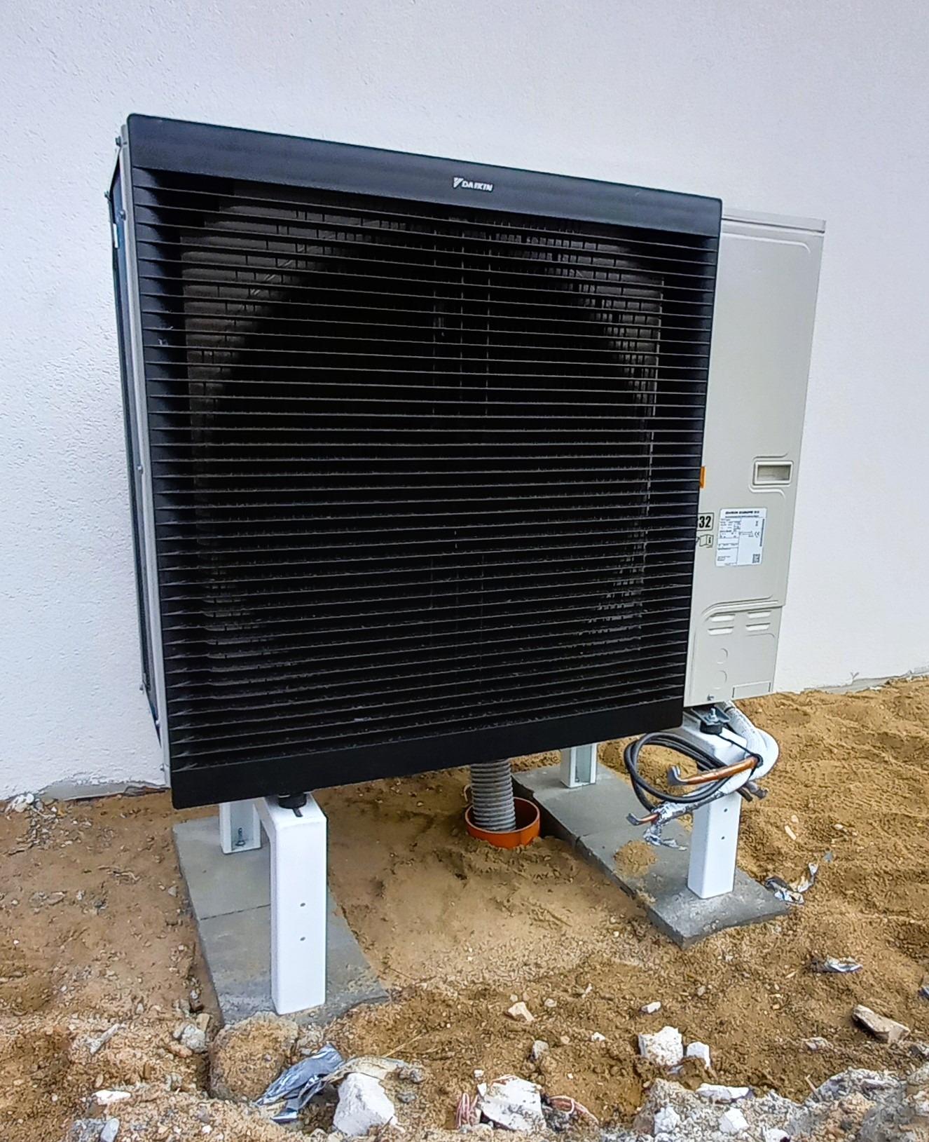 Zewnętrzna jednostka pompy ciepła Daikin na betonowych podstawach w trakcie instalacji, widoczne rury i kable, ziemia wokół.