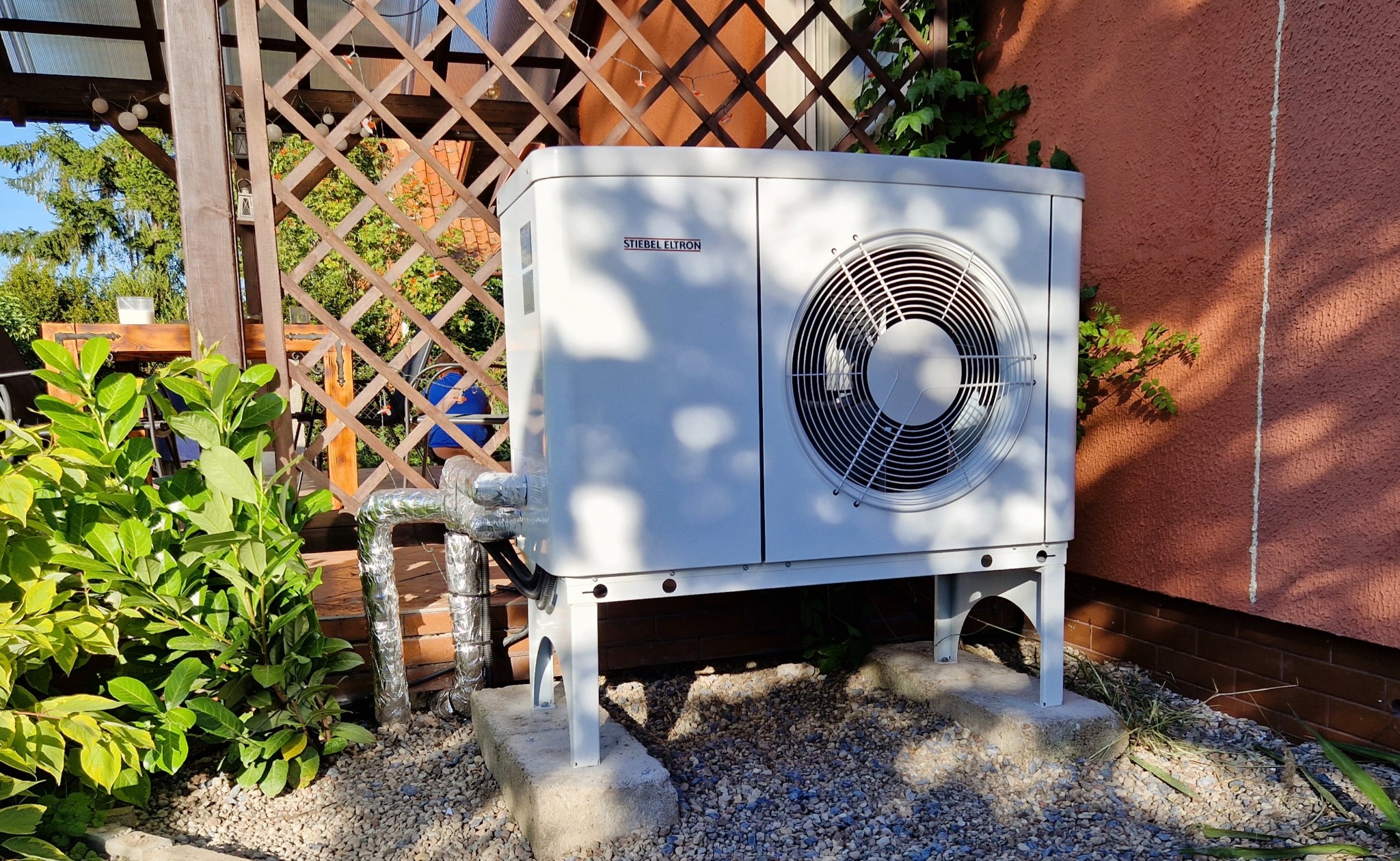 Biała pompa ciepła Stiebel Eltron zamontowana na zewnątrz budynku, widoczne rury w izolacji, tło z drewnianej kratownicy i zieleni.