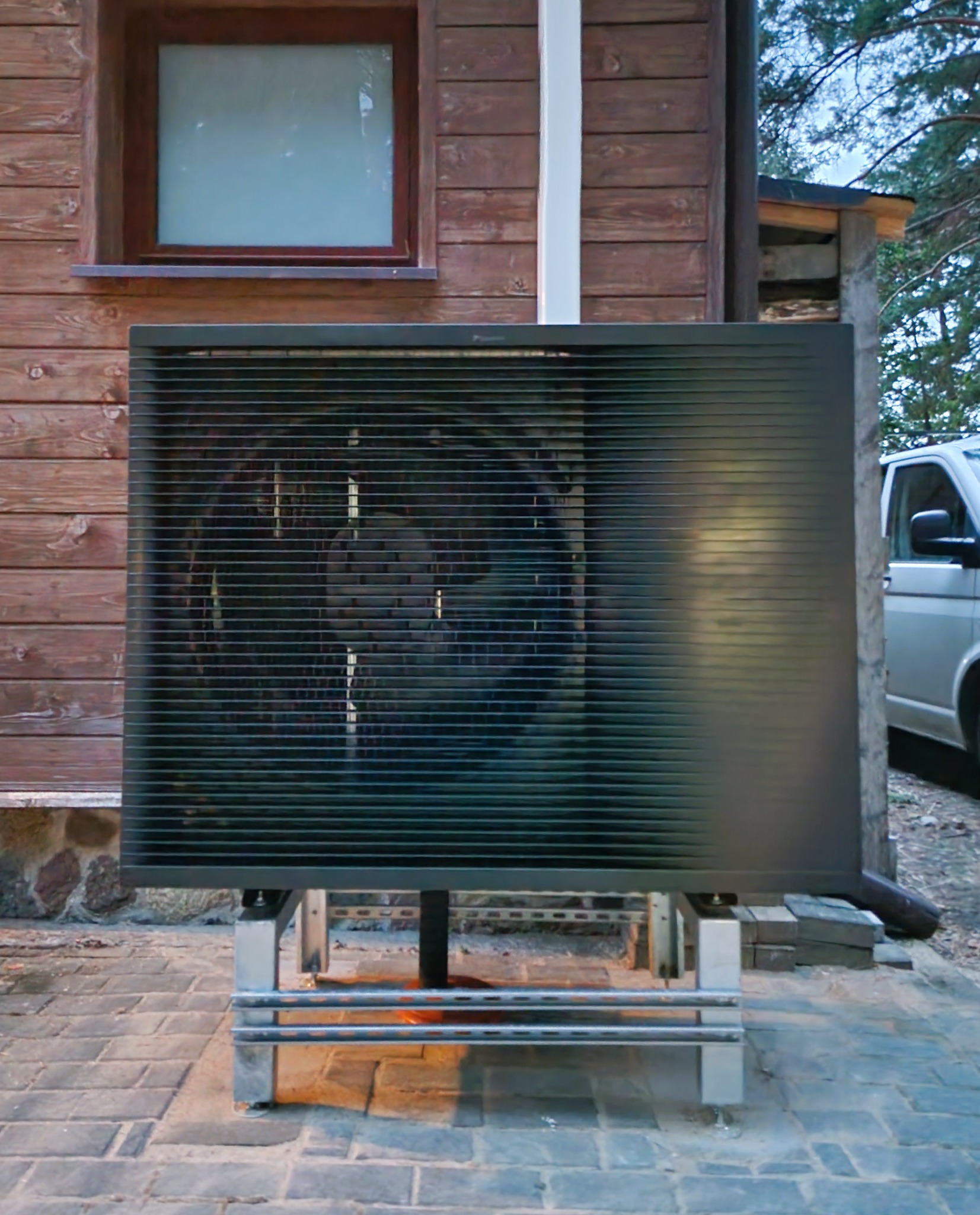 Zewnętrzna jednostka pompy ciepła Daikin zamontowana na metalowej konstrukcji na tle drewnianej ściany budynku z oknem, widoczny fragment zaparkowanego samochodu.