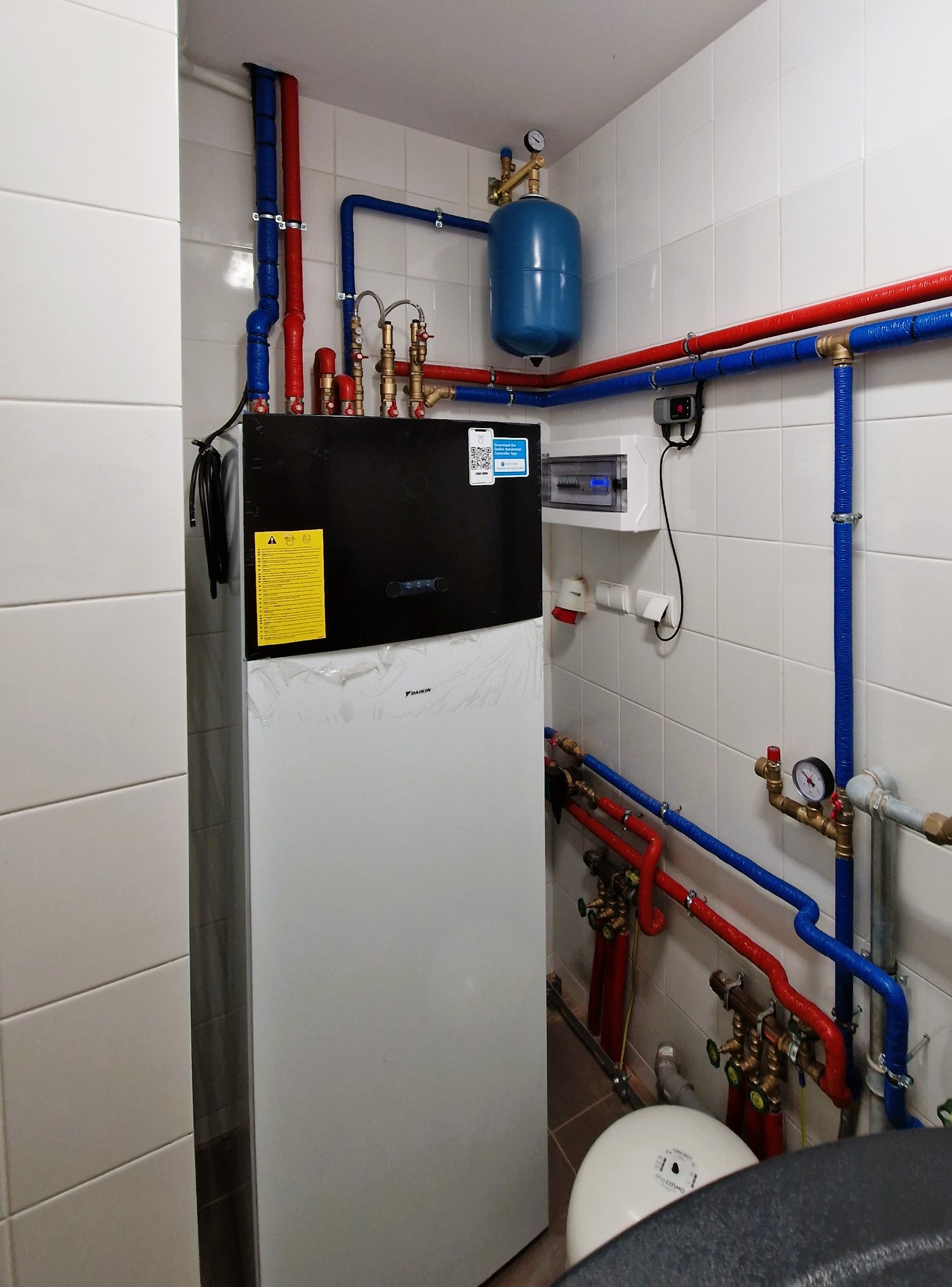 Nowoczesna instalacja pompy ciepła Daikin w kotłowni z białymi płytkami, widoczne rury w izolacji czerwonej i niebieskiej, manometry, zbiornik wyrównawczy i skrzynka elektryczna.