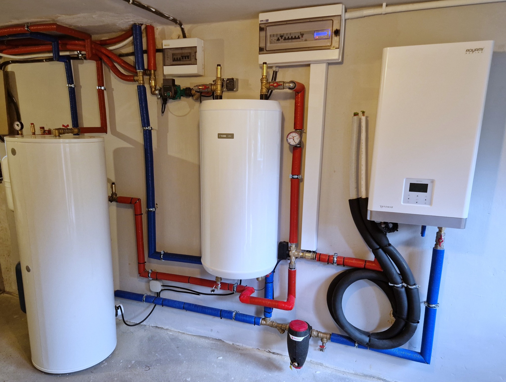 Instalacja kotła grzewczego i zasobnika ciepłej wody użytkowej w Pieniężnie, z widocznymi rurami w izolacji czerwonej i niebieskiej oraz elementami sterowania elektrycznego na ścianie.