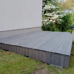 Drewniany taras w odcieniach szarości, przylegający do ściany domu, otoczony zielonym trawnikiem i kwitnącymi drzewami w tle. Na tarasie leży żółty nóż introligatorski.