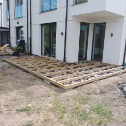 Drewniana konstrukcja tarasu na gruncie, przygotowana pod deski, z widocznymi kamieniami i geowłókniną, na tle nowoczesnego domu z dużymi oknami.