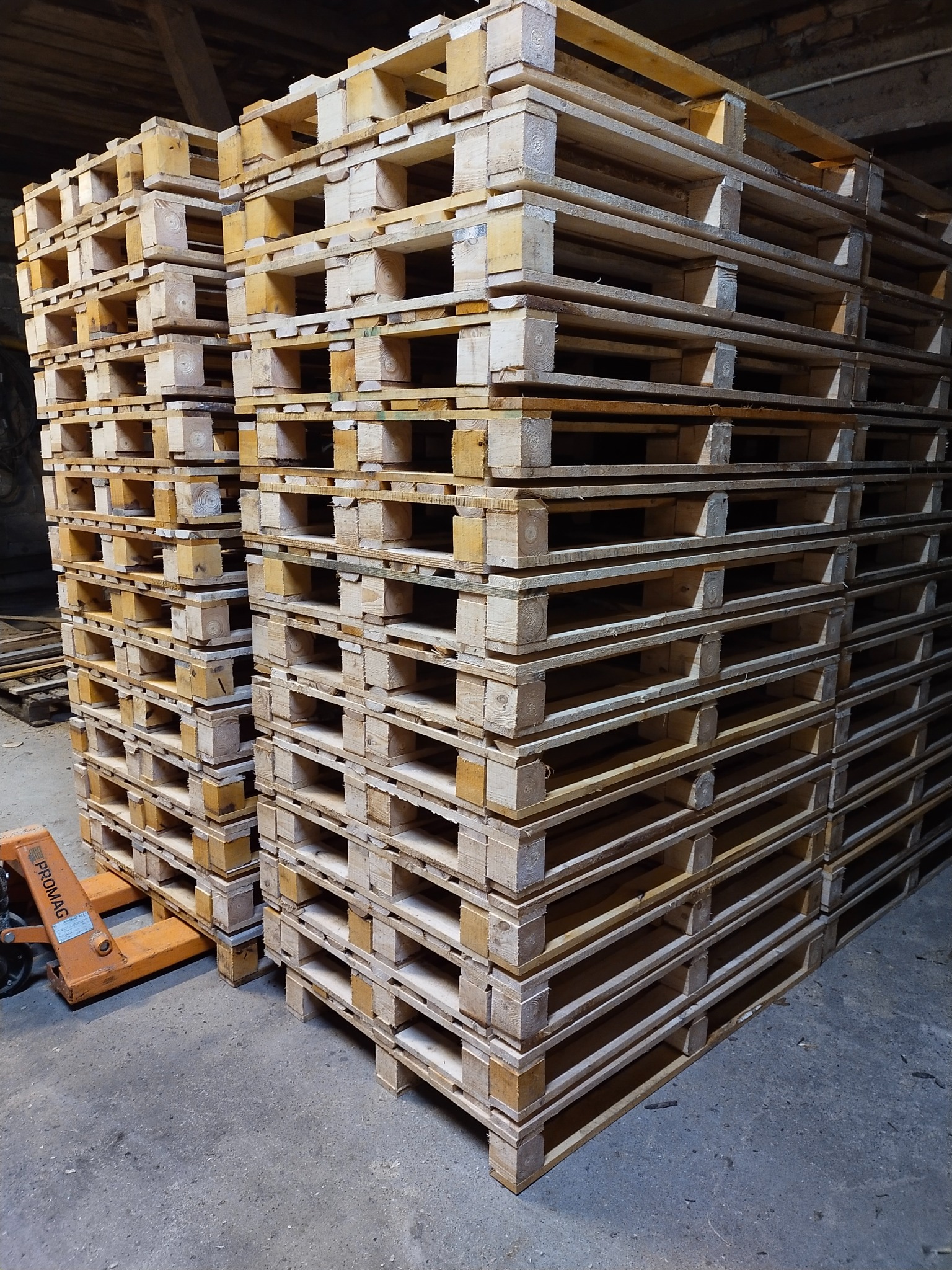 Dwa stosy drewnianych palet euro ułożone w magazynie, obok widoczny pomarańczowy wózek paletowy PROMAG.