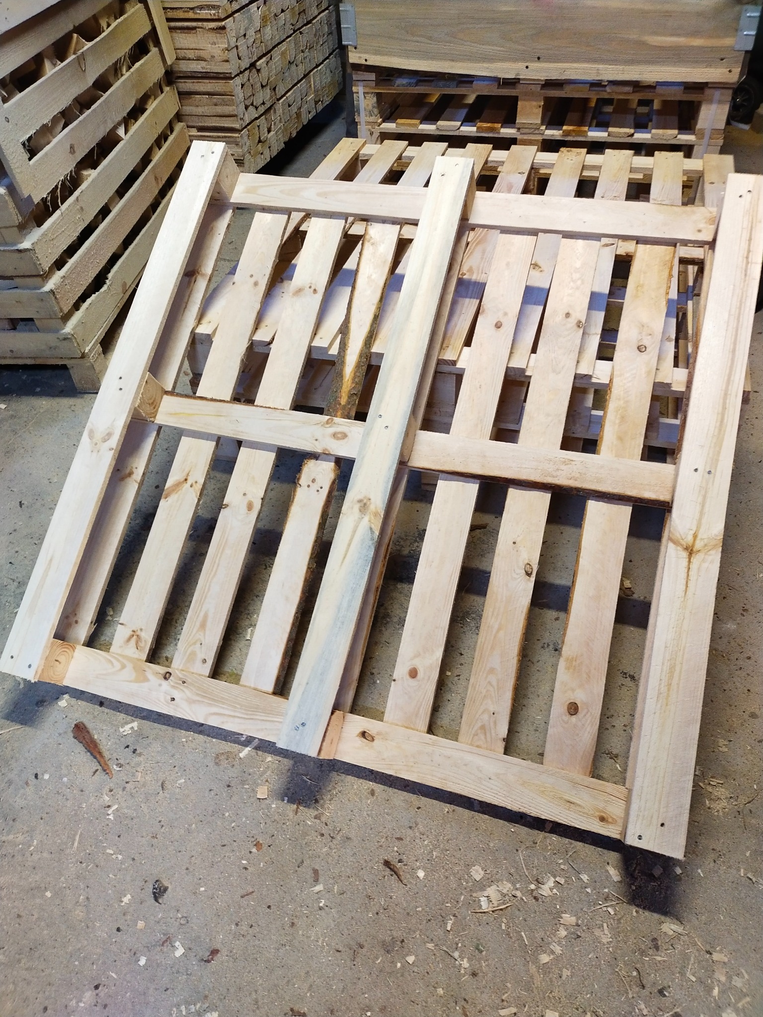 Jasne, drewniane palety ułożone jedna na drugiej, widoczne ślady po gwoździach i naturalne słoje drewna, na szarym, betonowym podłożu z drobnymi wiórami.