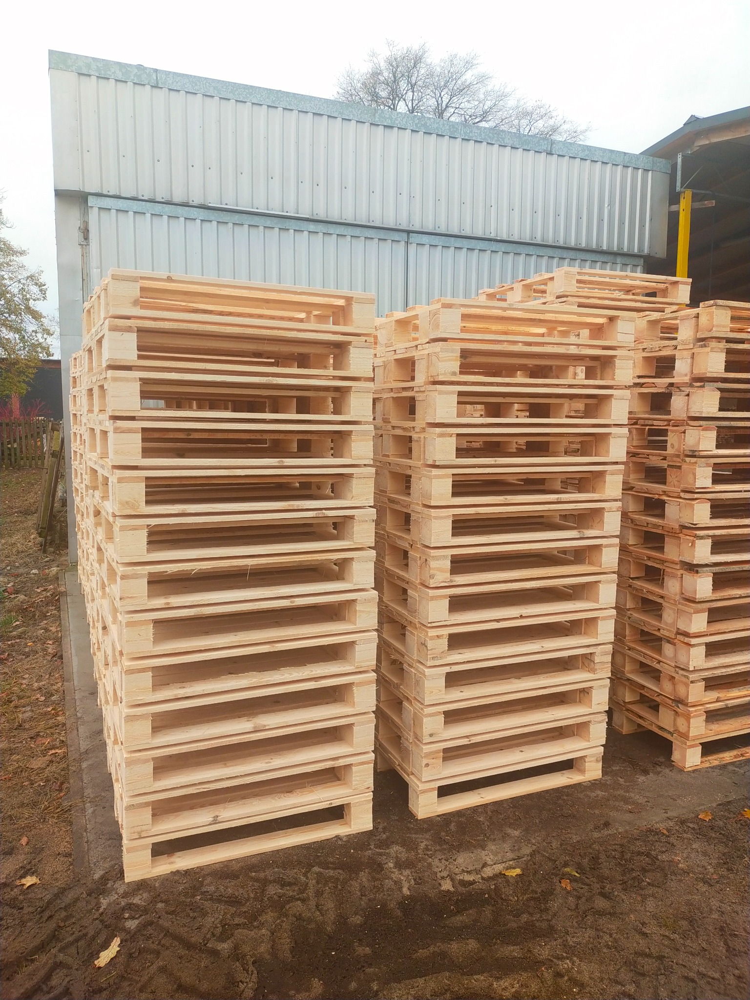 Stosy jasnych, drewnianych palet euro ułożone na zewnątrz, przed budynkiem z blachy falistej w pochmurny dzień.