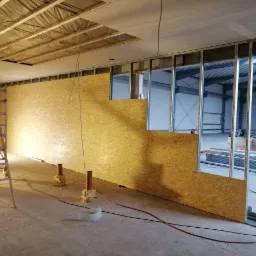 Montaż ścian działowych z płyty OSB na metalowym stelażu w hali, widoczne przewody elektryczne na podłodze i drabina aluminiowa