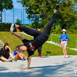 Dziewczyna w czarnym stroju robiąca stójkę na rękach podczas obozu tanecznego ViaCamp.pl, w tle grupa młodzieży i zielona trawa.
