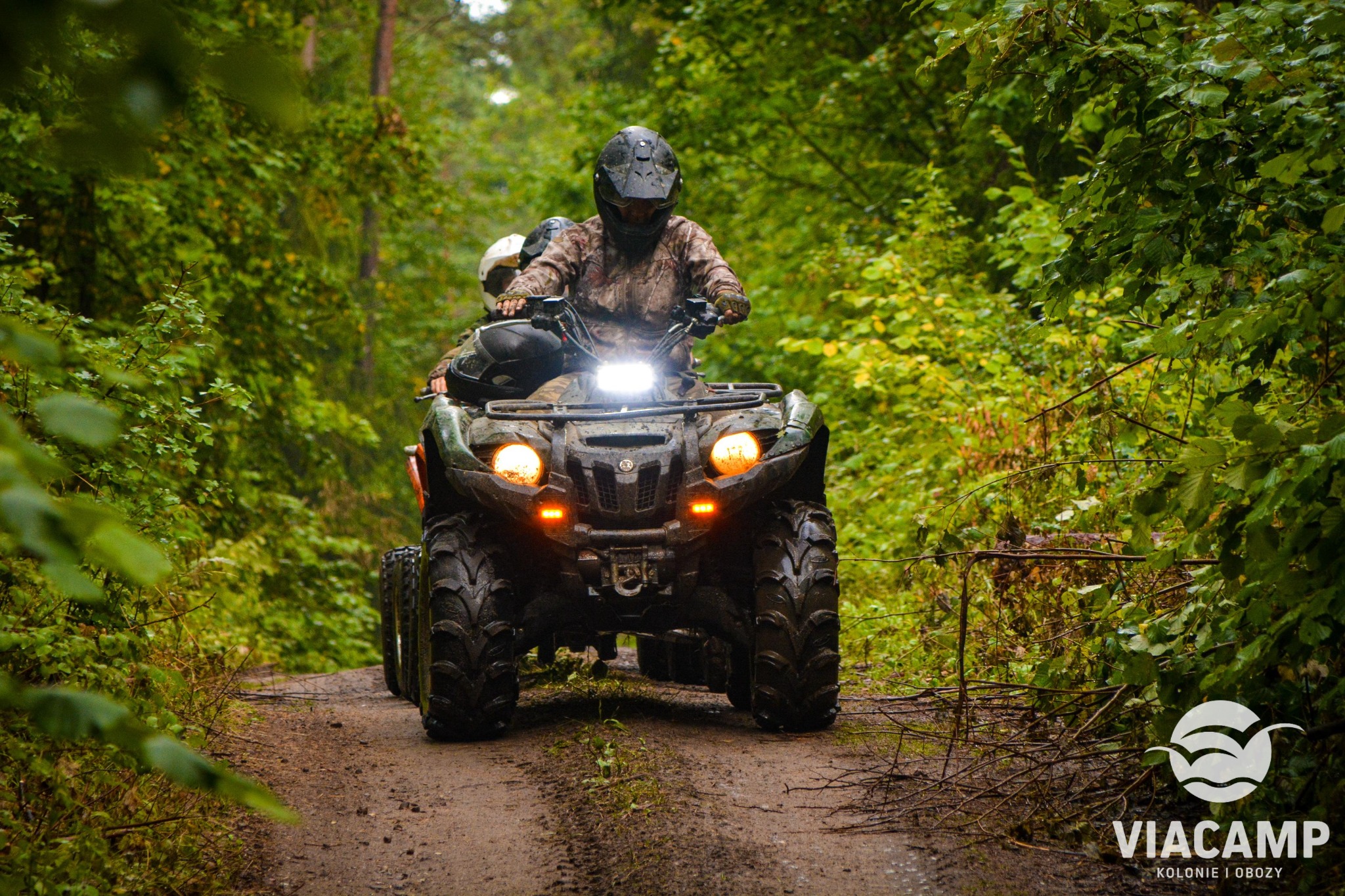 Dwóch uczestników w kaskach na quadzie jadących po błotnistej leśnej drodze, oświetlonych światłami pojazdu.