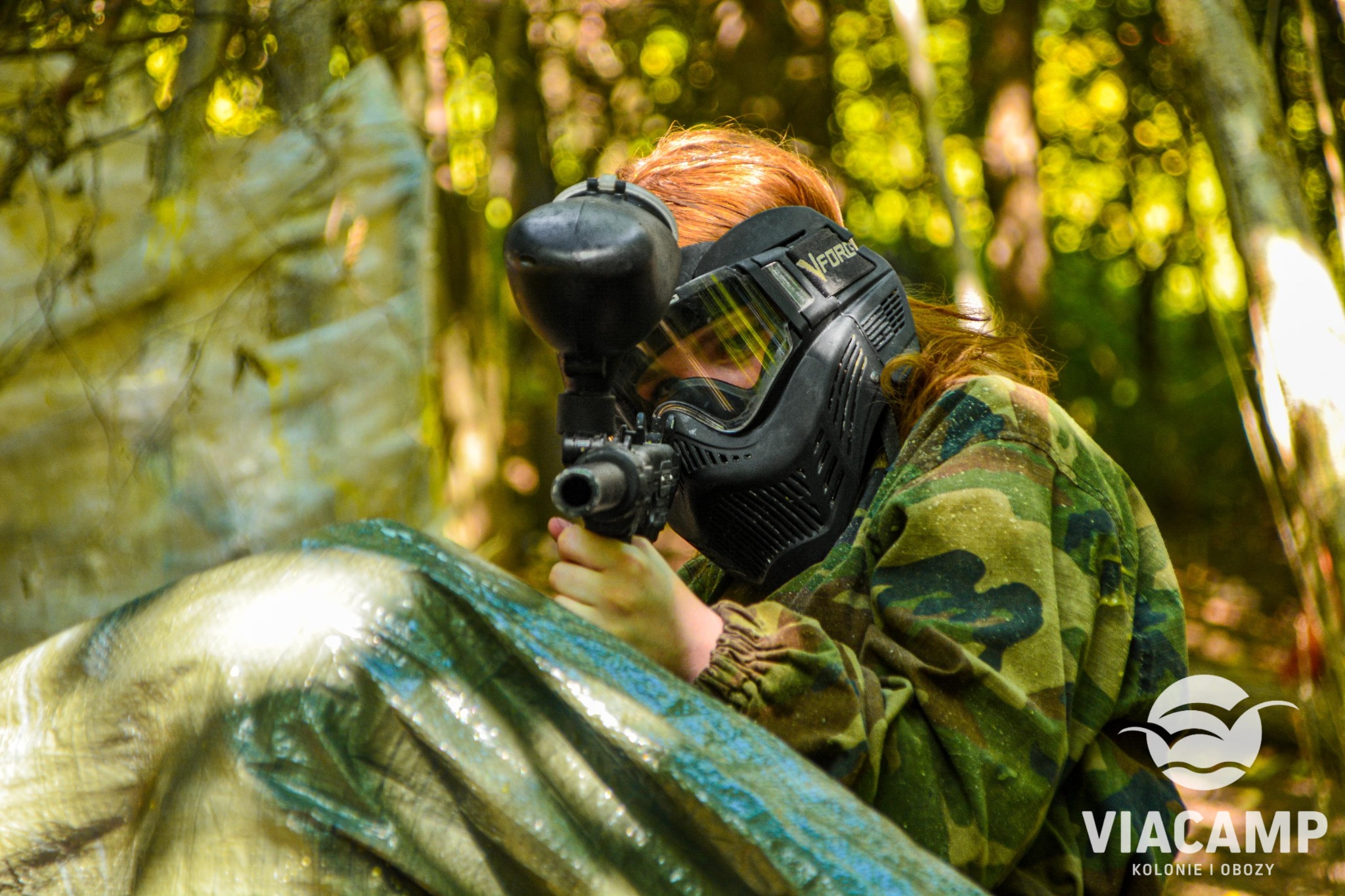 Osoba w kamuflażu i masce ochronnej celuje z markera paintballowego zza osłony, w tle rozmyty las.
