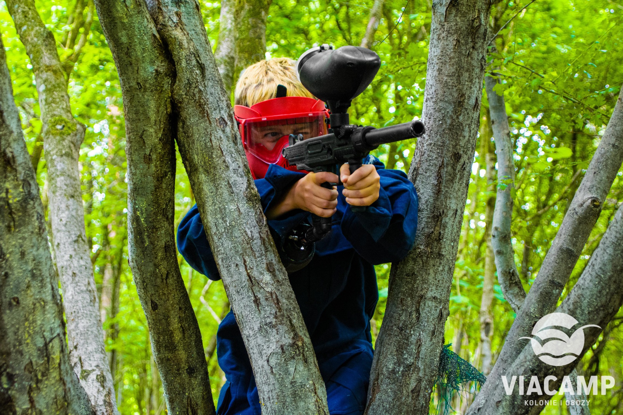 Chłopiec w czerwonej masce ochronnej i niebieskim kombinezonie celuje z karabinu paintballowego, ukryty za drzewami w lesie.