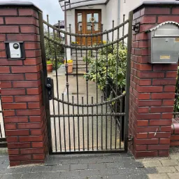 Kuta brama wjazdowa z metalowymi prętami i ceglanymi słupkami w Obornikach, z widoczną skrzynką na listy i domofonem.