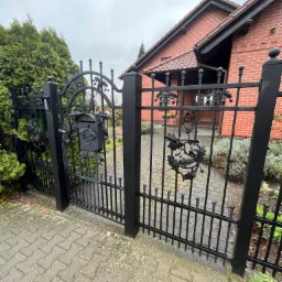 Czarna, kuta brama z motywem liści dębu i żołędzi, zintegrowana ze skrzynką na listy, prowadząca do domu z czerwonej cegły w pochmurny dzień.