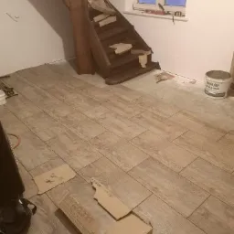 Układanie płytek imitujących drewno na podłodze, widoczne fragmenty schodów, okno z widokiem na zaśnieżony krajobraz i wiadro z zaprawą klejową