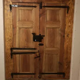 Odrestaurowane, drewniane drzwi z czarnymi, kutymi zawiasami i zamkiem, osadzone w surowej ościeżnicy z płyt gipsowo-kartonowych, widoczne ślady renowacji i starej struktury drewna.