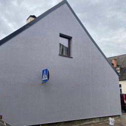 Dk trans holding Sp.zo.o. - Świeżo pomalowana na szaro ściana budynku z małym oknem i znakiem przejścia dla pieszych, widoczna drabina i wiadro z farbą przy chodniku.