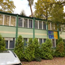 Dk trans holding Sp.zo.o. - Dwukondygnacyjny budynek z zieloną elewacją z poziomych paneli, widoczne drzewa i krzewy iglaste, na oknie kartka z napisem 'Sprzedam' i numerem telefonu, częściowo zasłonięty przez dach samochodu.