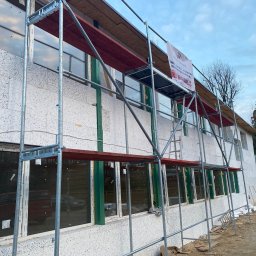 Dk trans holding Sp.zo.o. - Budynek dwukondygnacyjny w trakcie ocieplania, widoczne rusztowanie, styropian na ścianach, okna z zielonymi ramami, baner reklamowy firmy budowlanej na rusztowaniu.
