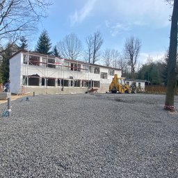 Dk trans holding Sp.zo.o. - Dwukondygnacyjny budynek w trakcie ocieplania elewacji, widoczne rusztowania, mieszalnik betonu i koparka na placu budowy z wysypanym szarym gruzem.