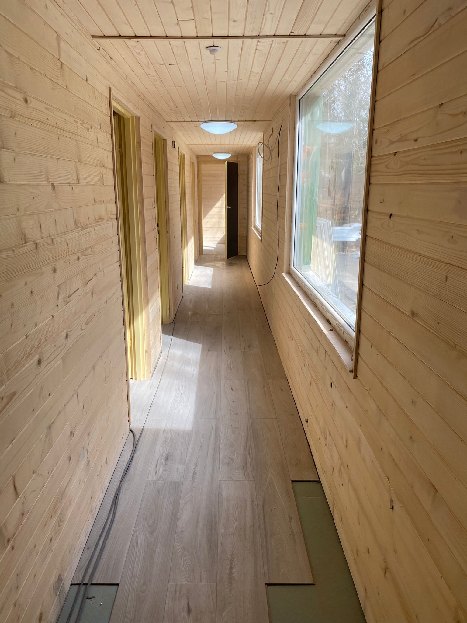 Korytarz wyłożony boazerią z jasnego drewna, z widocznymi otworami drzwiowymi i dużym oknem, podłoga częściowo wyłożona panelami, widoczne przewody elektryczne przy ścianie.