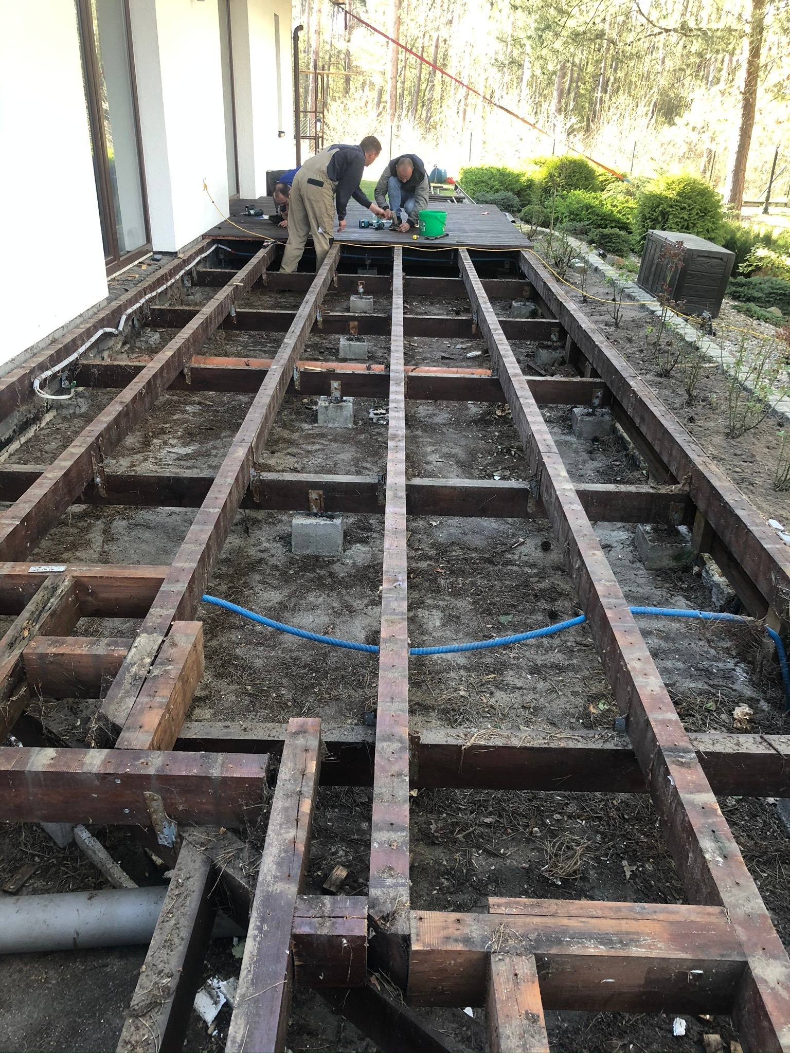 Montaż drewnianej konstrukcji tarasu na betonowych wspornikach przy budynku, widoczne przewody elektryczne i hydrauliczne, dwóch pracowników w roboczych ubraniach przykręca deski.