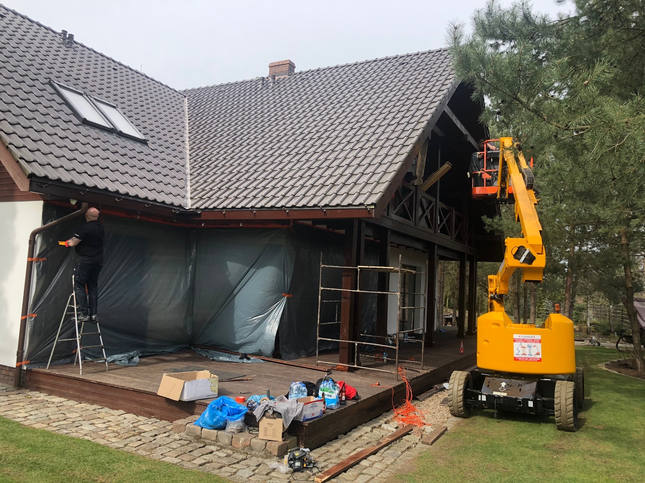 Prace na elewacji domu jednorodzinnego z wykorzystaniem podnośnika koszowego i rusztowań, zabezpieczone folią, widoczny pracownik na drabinie.