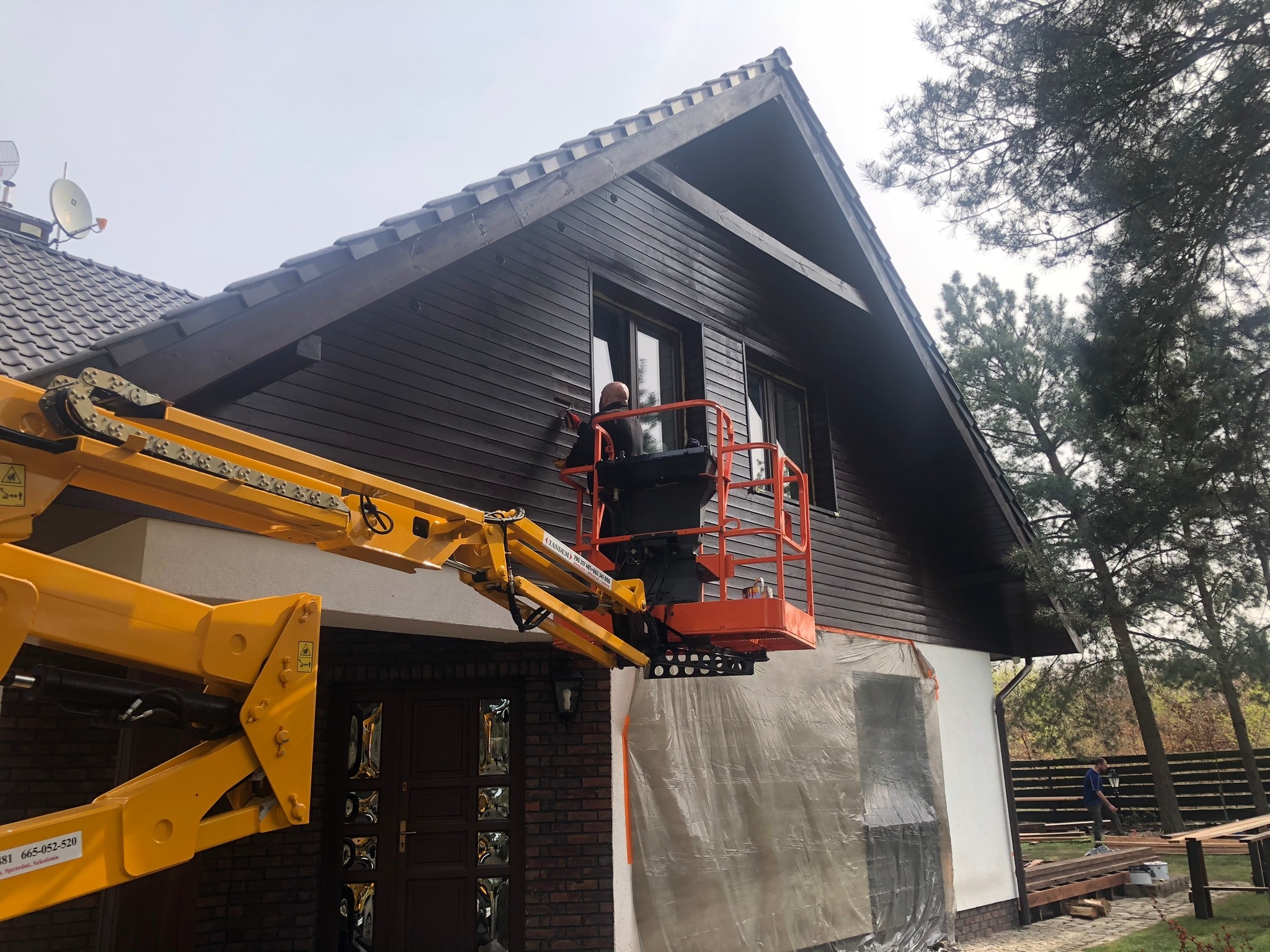 Pracownik maluje drewnianą elewację domu z podnośnika koszowego, zabezpieczona folią część ściany, widoczne drzewa w tle.